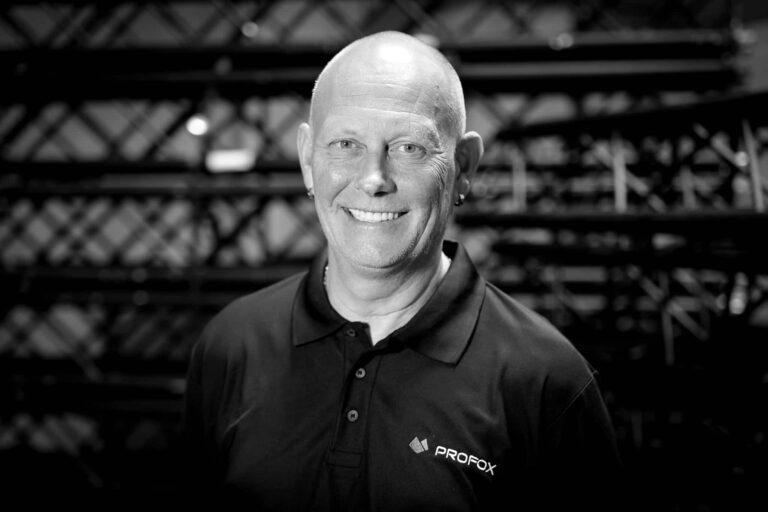 A smiling bald man wearing a dark Profox polo shirt stands in front of a background with metal trusses, photographed in black and white.