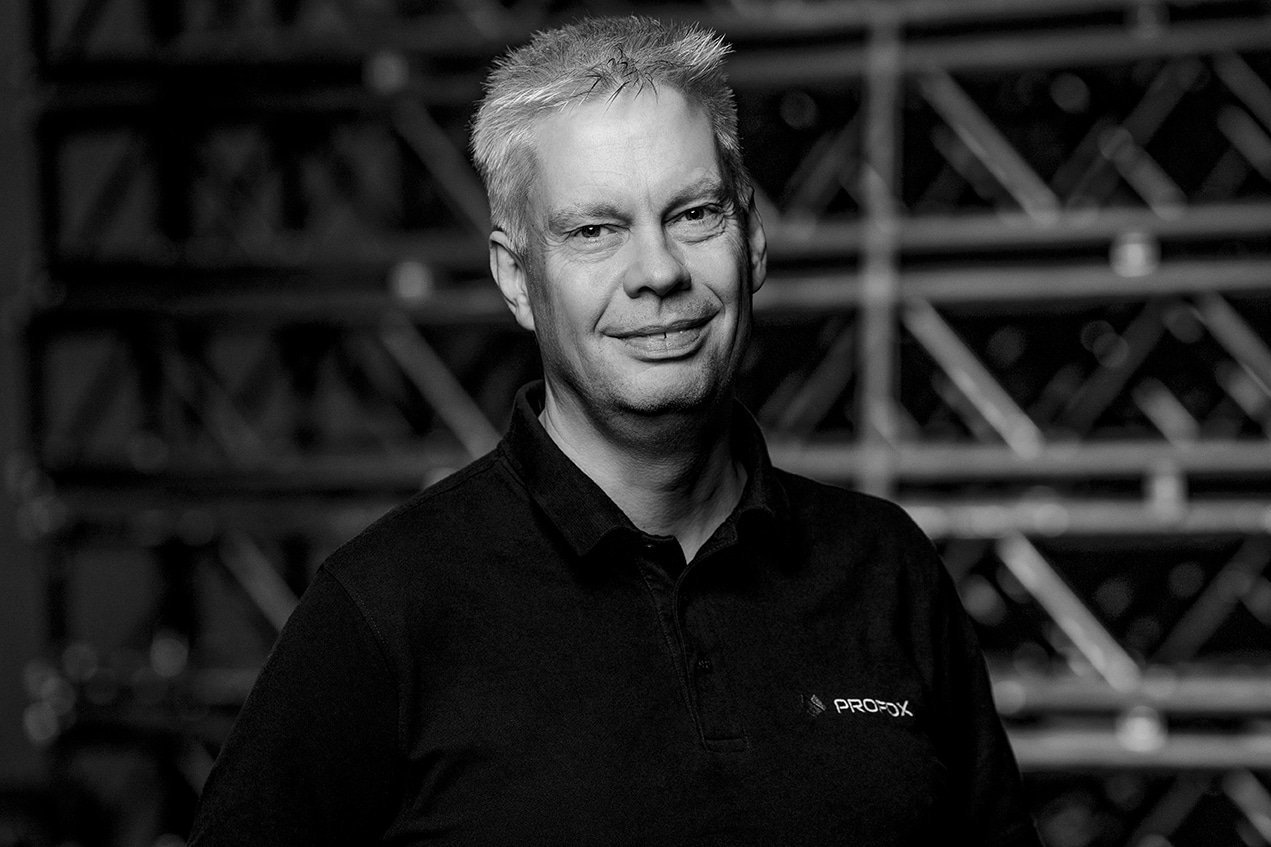 A man with short, light hair wearing a dark polo shirt stands in front of metal shelving. The image is in black and white, and the man is smiling slightly while looking at the camera.