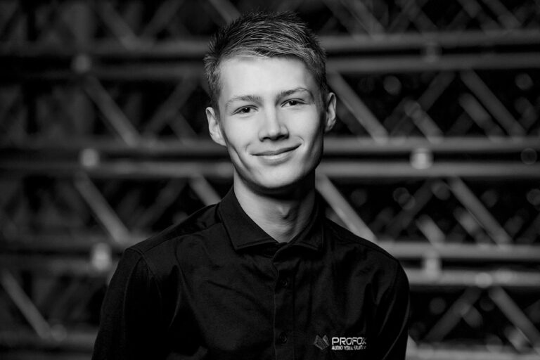 A young man with short hair smiles at the camera, wearing a dark collared shirt with a PROFOX AUDIO VISUAL logo. The background features out-of-focus, crisscrossed metal structures. The image is in black and white.