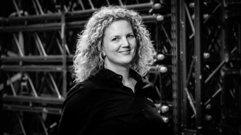 A woman with curly hair, wearing a dark shirt, smiles at the camera while standing in front of a metal structure. The image is in black and white.