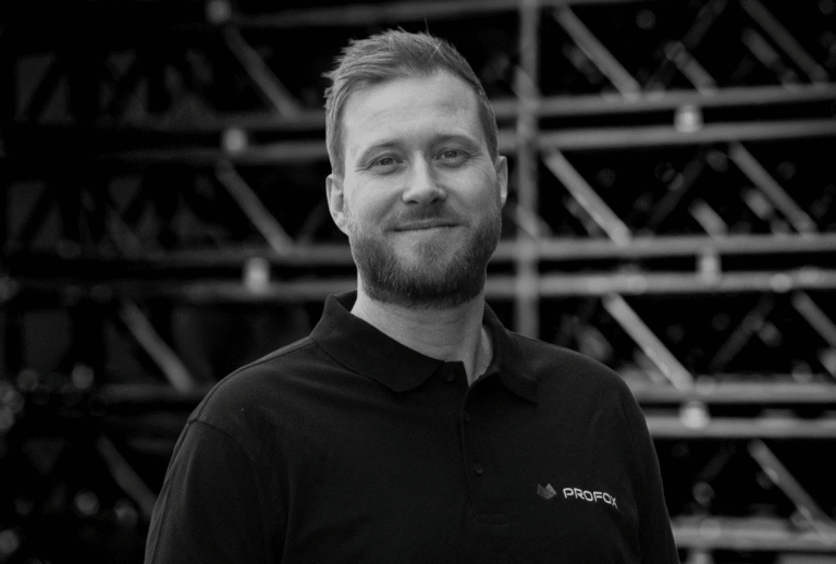 A man with short hair and a beard, wearing a black collared shirt with a PROFOL logo, stands smiling in front of a background filled with metal beams. The image is in black and white.