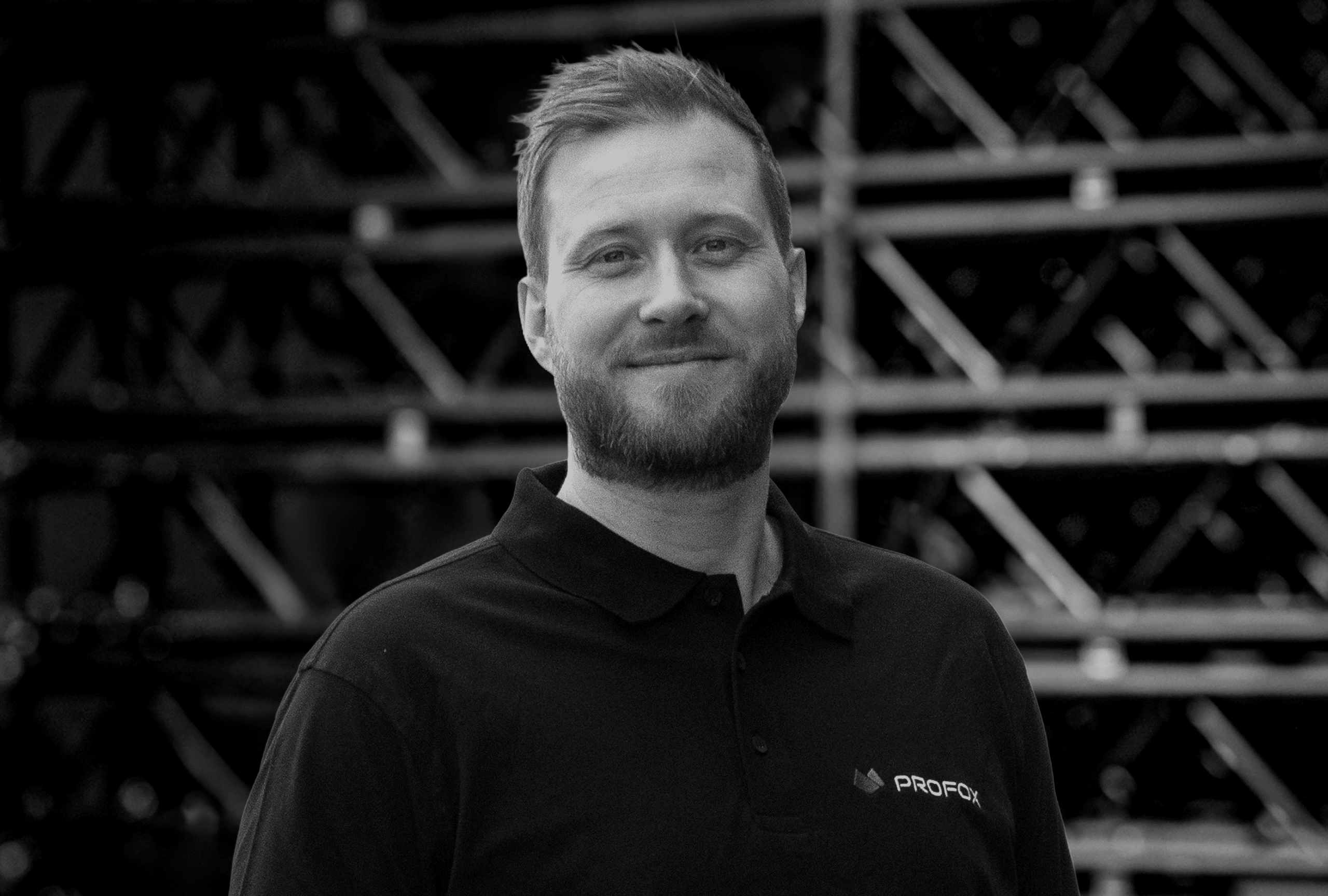 A man with short hair and a beard, wearing a black collared shirt with a PROFOL logo, stands smiling in front of a background filled with metal beams. The image is in black and white.