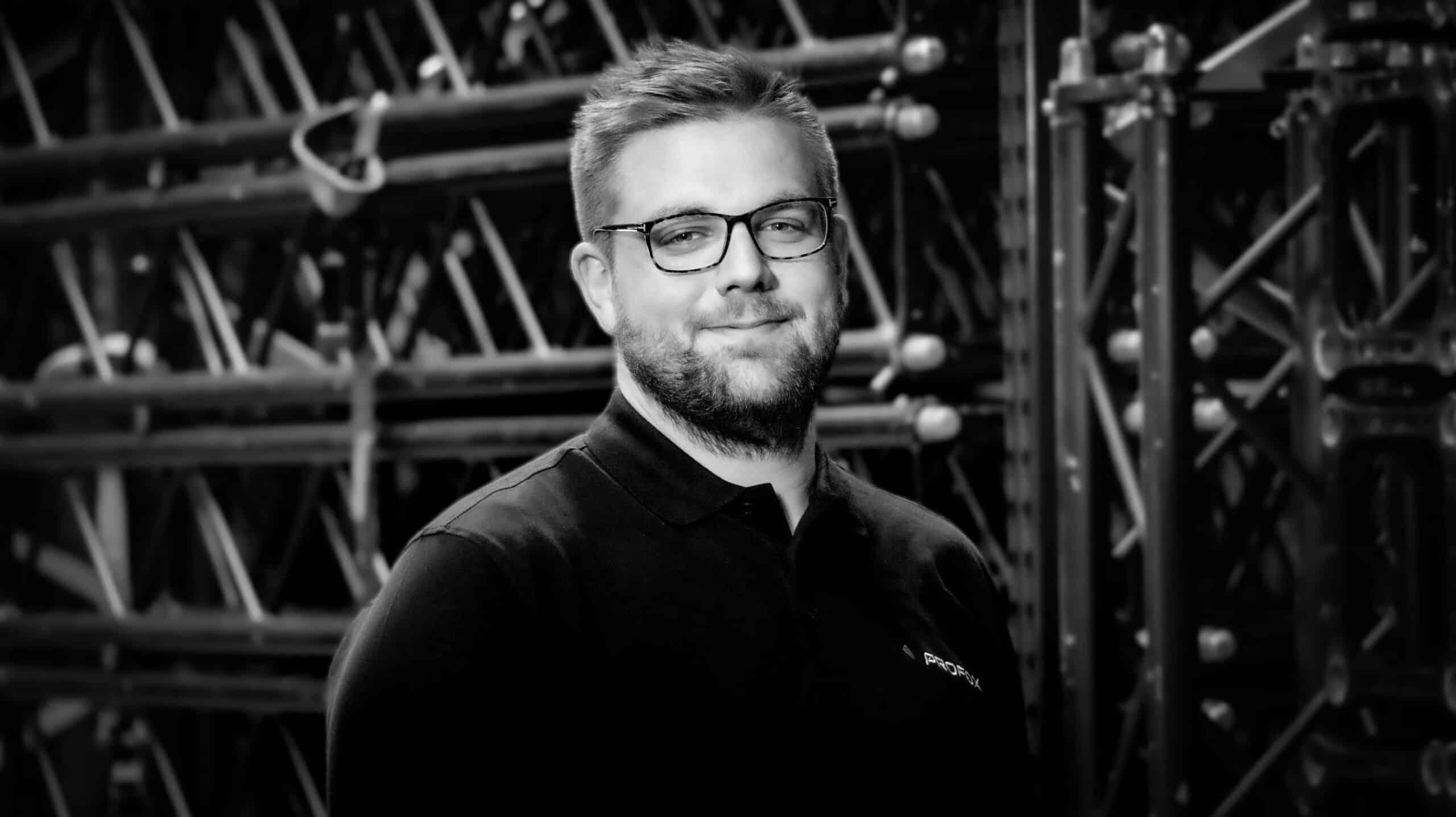 A man with short hair, glasses, and a beard, wearing a dark polo shirt, stands in front of a background filled with metal scaffolding or equipment. The image is black and white.