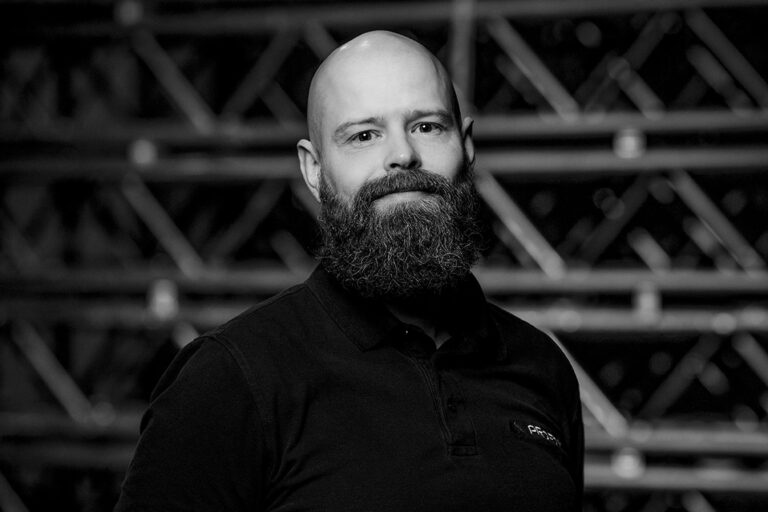 A bald man with a thick, full beard wears a dark polo shirt and stands in front of a background of metal scaffolding. The photo is black and white, and he is looking at the camera with a slight smile.