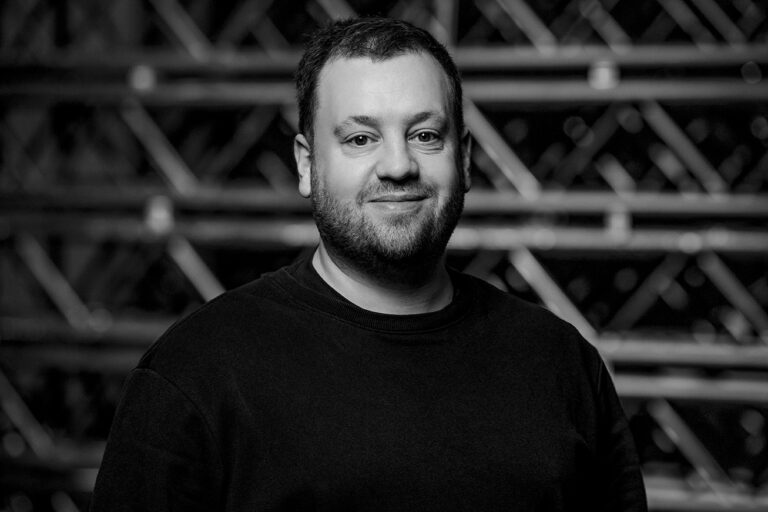A man with short hair and a beard, wearing a dark sweater, smiles slightly while standing in front of an out-of-focus industrial or metal background. The photo is in black and white.