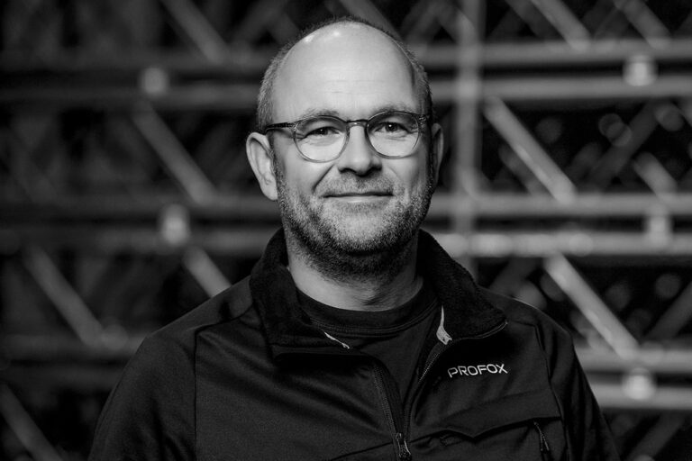 A man with glasses and a beard, wearing a dark jacket with the word PROFOX on it, smiles slightly while standing in front of a blurred industrial background. The photo is in black and white.