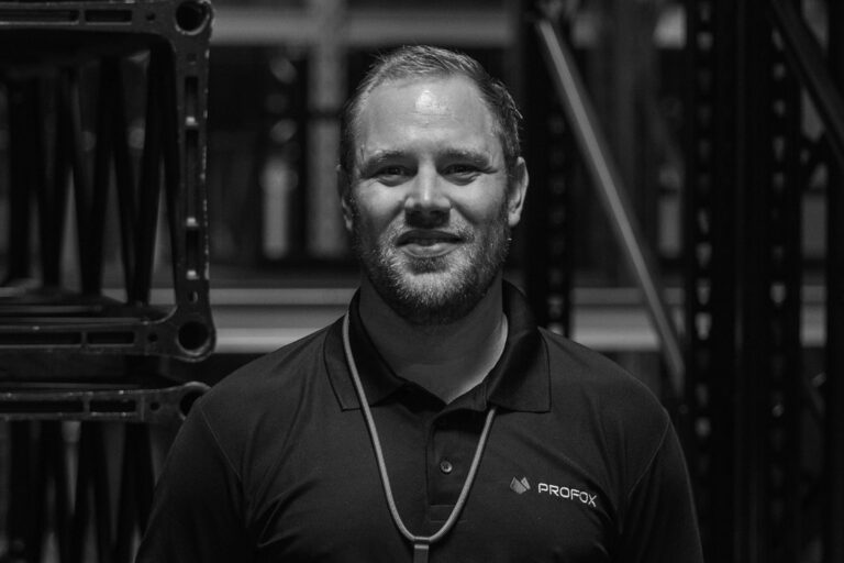 A man with a beard, wearing a dark polo shirt with a PROFOX logo and a lanyard, stands in front of industrial shelving in a warehouse. The image is in black and white.