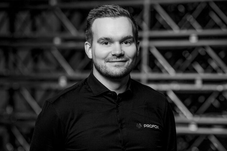 A man with short hair and a beard, wearing a dark dress shirt with the logo “PROFOX,” smiles at the camera. The background is out of focus, featuring metal scaffolding or racks. The image is in black and white.