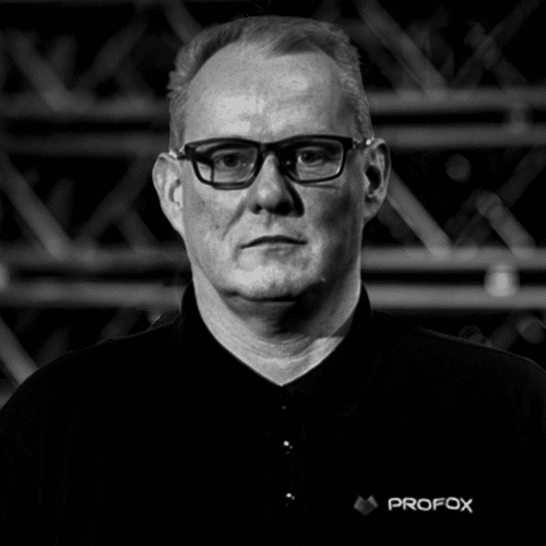 A middle-aged person with short light hair and glasses, wearing a dark collared shirt with a PROFOX logo, stands in front of a blurred industrial background.