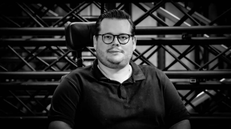 A man with glasses and short hair, wearing a dark polo shirt, sits in a wheelchair and faces the camera. The background features geometric metal bars. The photo is in black and white.
