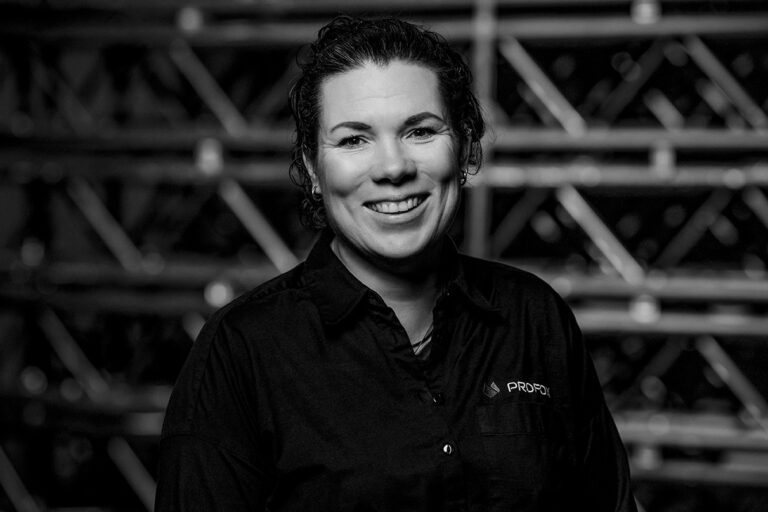 A woman with curly hair, wearing a dark shirt and smiling, stands in front of a blurred background featuring metal racks. The image is in black and white.