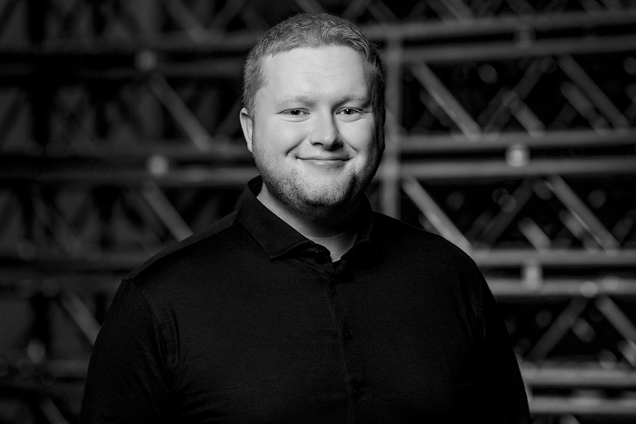 A man with short hair and a beard, wearing a dark collared shirt, smiles at the camera. The background is filled with crisscrossed metal beams. The photo is in black and white.