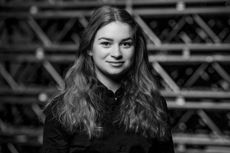 A young woman with long wavy hair and a slight smile, wearing a dark shirt, stands in front of a blurred background with intersecting lines. The image is in black and white.