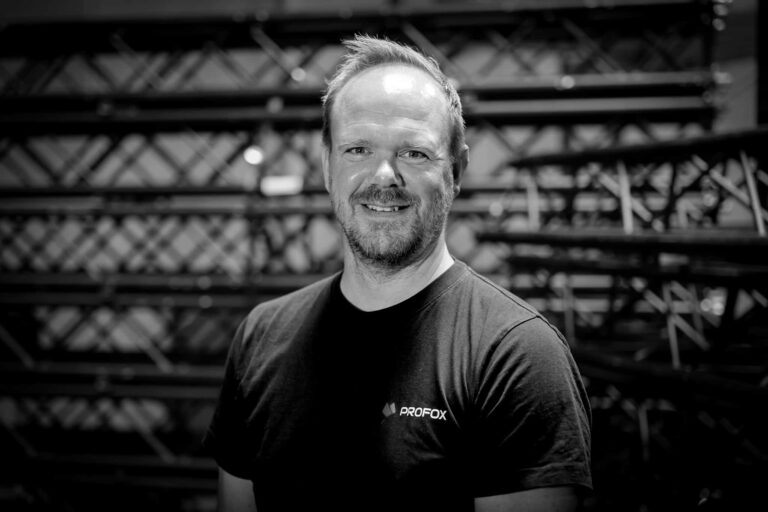 A man with short hair and a beard, wearing a dark PROF0X t-shirt, smiles while standing in front of stacked metal beams. The image is in black and white.