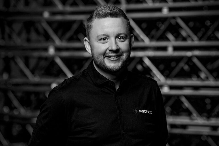 A man with short hair and a beard smiles at the camera, wearing a dark button-up shirt with a logo that reads PROFCK. The background features a pattern of metal bars and beams. The image is in black and white.