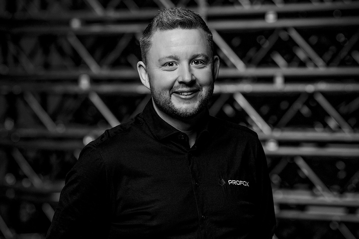 A man with short hair and a beard smiles at the camera, wearing a dark button-up shirt with a logo that reads PROFCK. The background features a pattern of metal bars and beams. The image is in black and white.