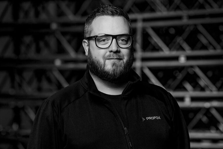 A bearded man wearing glasses and a dark jacket with a PROFEX logo stands in front of a blurred industrial background, looking at the camera and smiling slightly. The image is black and white.