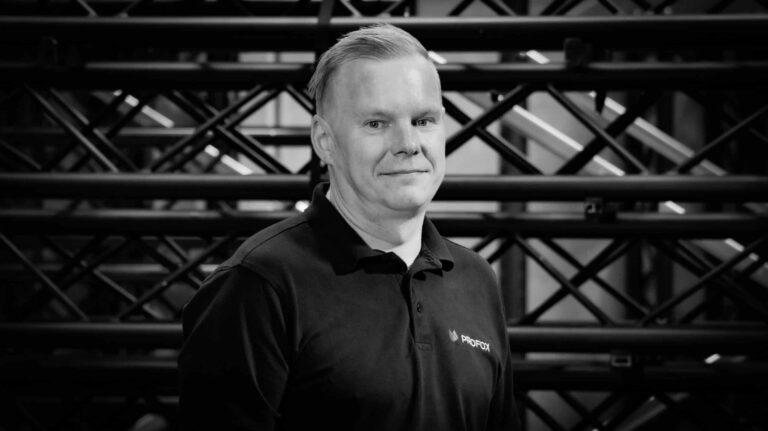 A man with short light hair wearing a dark polo shirt stands in front of a background of metal beams or scaffolding, looking at the camera and smiling slightly. The image is in black and white.