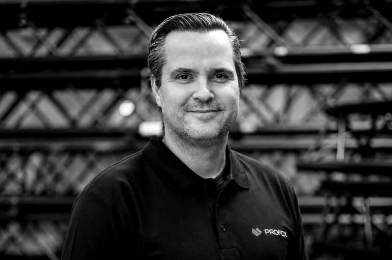 A man with short, neatly combed hair and light stubble smiles at the camera, wearing a black polo shirt with a logo. The background features blurred industrial-style metal racks.