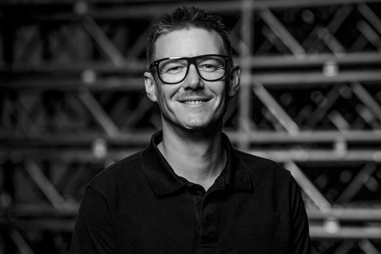 A smiling person with short hair, wearing large glasses and a dark polo shirt, stands in front of an industrial-style background with metal beams. The image is in black and white.
