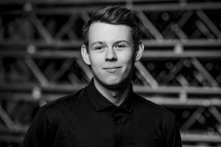 A young man in a dark, buttoned-up shirt stands in front of a blurred background with a faint grid pattern, looking slightly to the side and smiling softly. The image is black and white.