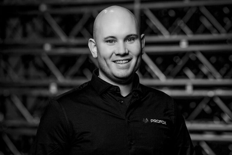 A smiling bald man in a dark collared shirt with a PROFOX logo, standing in front of a blurred background with diagonal lines. The image is in black and white.