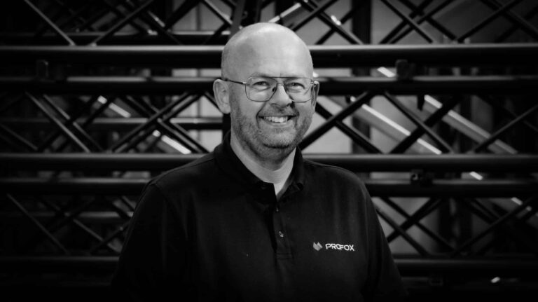A smiling man with glasses and a beard, wearing a dark polo shirt with the logo PROFOX, stands in front of a background of intersecting metal beams. The image is black and white.