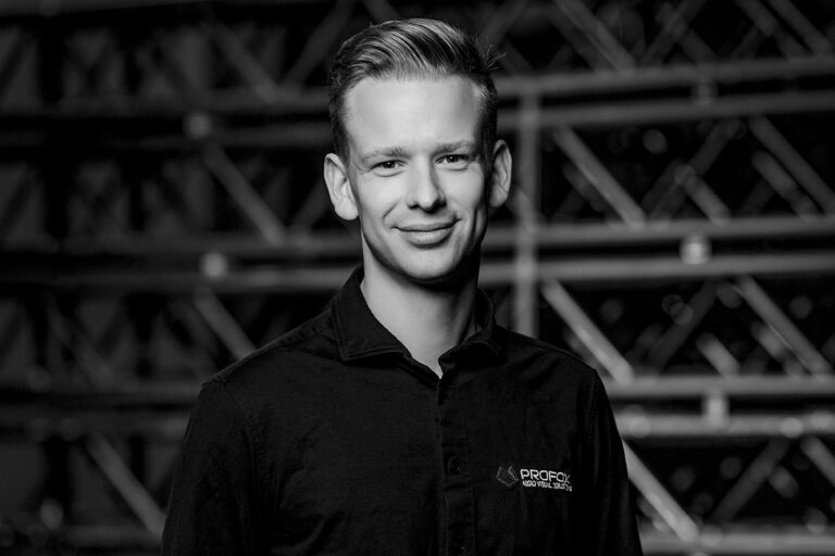 A man with short, styled hair wearing a dark collared shirt with a “PROFIC” logo, stands smiling in front of a background with diagonal metal beams. The image is in black and white.