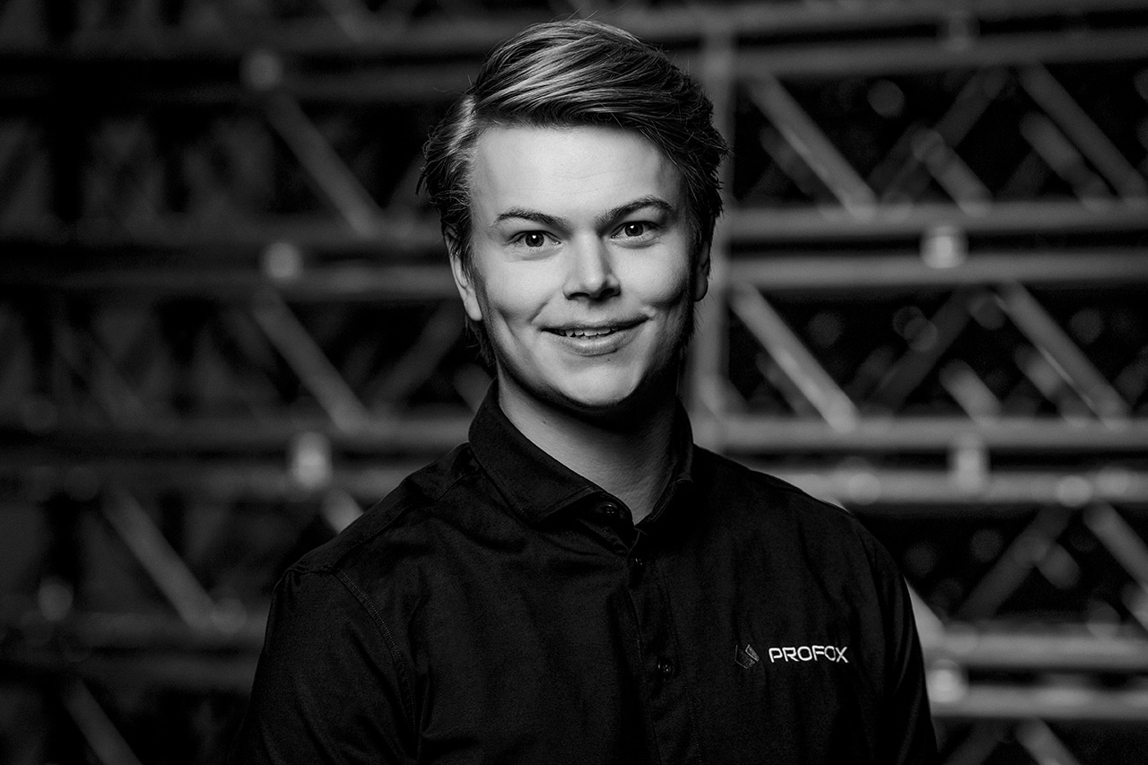 A young person with short, styled hair smiles at the camera, wearing a dark collared shirt with a PROFOX logo. The background is blurred, showing a geometric metal structure. The image is in black and white.