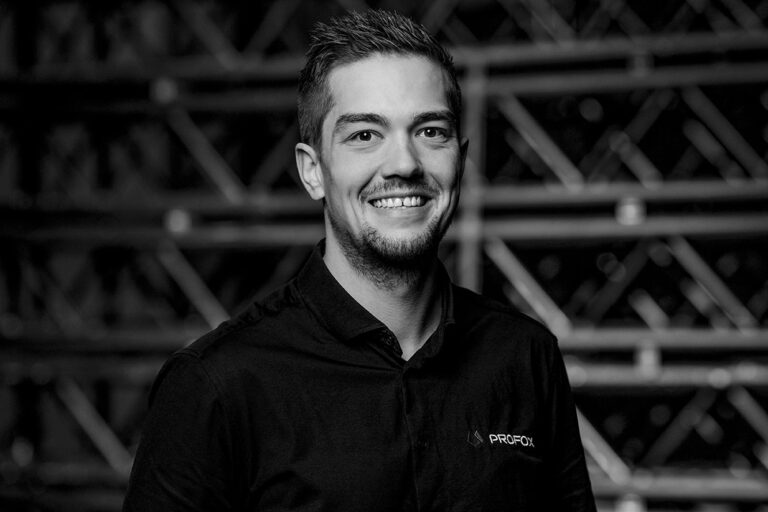 A young man with short hair and a trimmed beard smiles at the camera, wearing a dark button-up shirt with a PROFOX logo. The background features blurred industrial-style metal beams. The image is in black and white.
