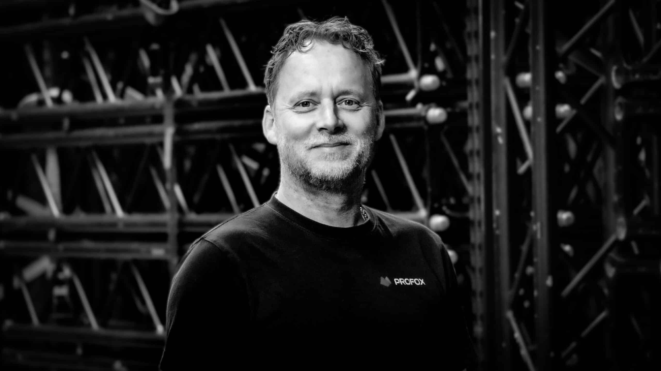 Black and white photo of a smiling man with short, curly hair and a light beard, wearing a dark t-shirt with a small logo, standing in front of a background with metal scaffolding.