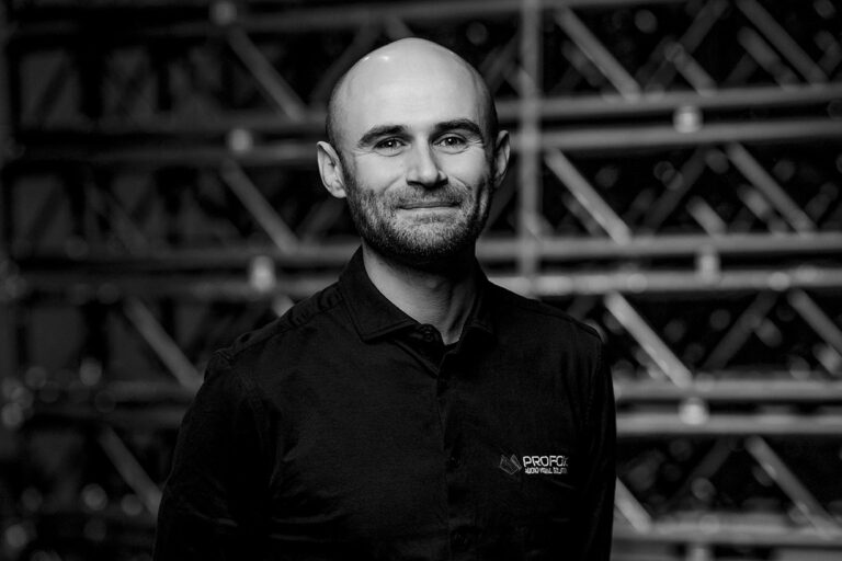 A man with a shaved head and beard smiles at the camera, wearing a dark collared shirt with a PROPC logo. The background features blurred metal structures, giving an industrial feel. The image is in black and white.