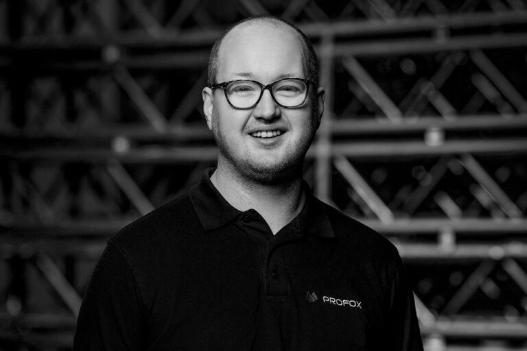 A man with glasses and short hair smiles at the camera, wearing a black collared shirt with the logo PROFOX on it. The background features an industrial-style metal framework, and the image is in black and white.