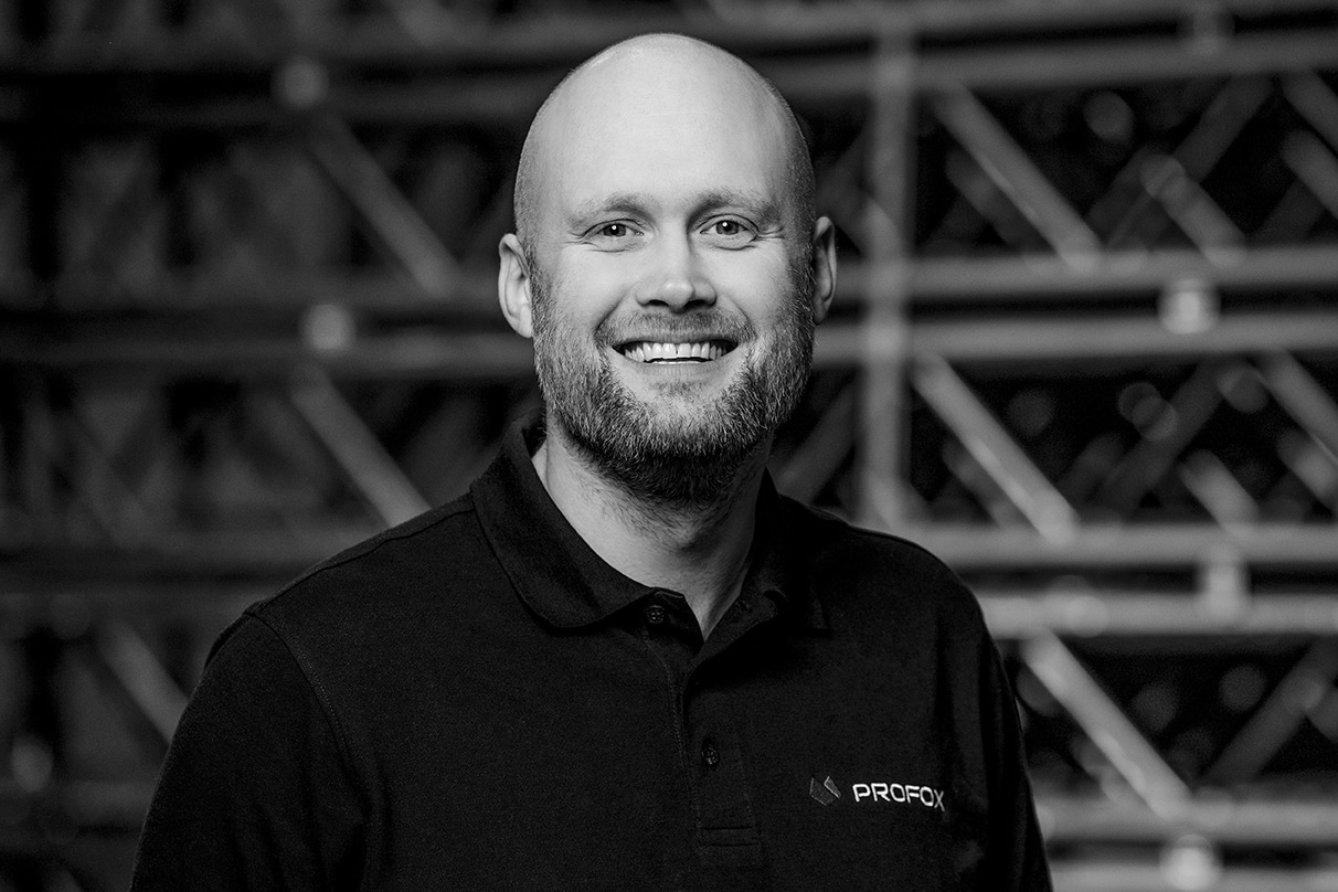 A smiling man with a bald head and beard wears a black polo shirt with a PROFOK logo. The background is out of focus, featuring geometric metal structures. The image is in black and white.