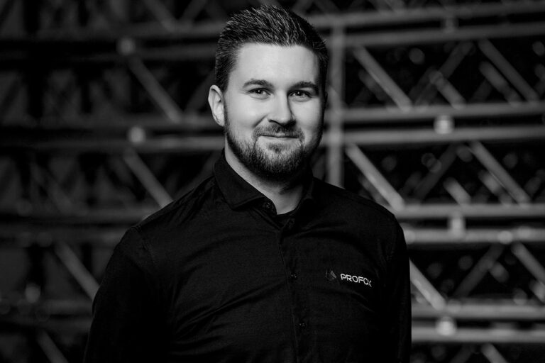 A man with short dark hair and a beard, wearing a black collared shirt with a small logo, stands and smiles at the camera. The background is blurred with a pattern of metal bars. The photo is in black and white.