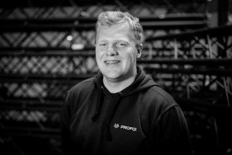 A smiling person with short light hair wearing a hoodie with the PROFOX logo, standing in front of a background featuring metal beams and trusses. The image is in black and white.