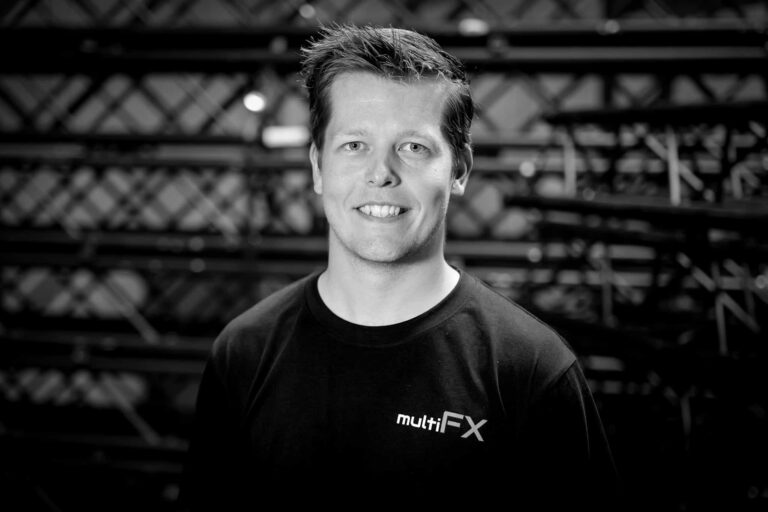 A smiling man wearing a multiFX t-shirt stands in front of metal scaffolding. The image is in black and white.
