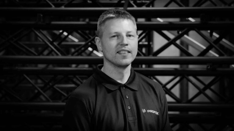 A man with short hair in a collared shirt stands in front of a metal or industrial geometric background, looking at the camera and smiling slightly. The image is in black and white.