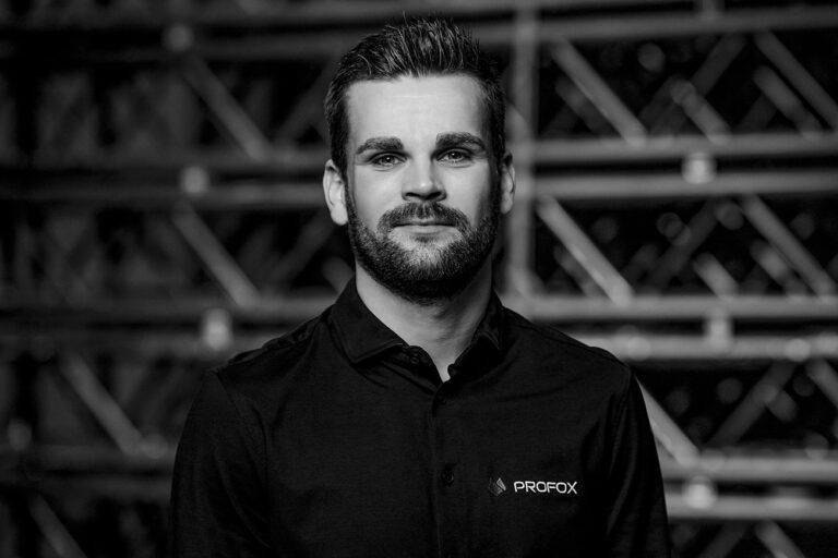 A man with short hair and a beard, wearing a dark collared shirt with a PROFOX logo, stands in front of a blurred industrial-style background. The image is in black and white.