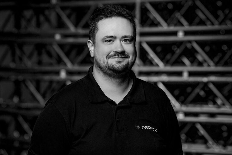 A man with short dark hair and a beard smiles while wearing a black polo shirt with a PRODIX logo. He stands in front of a blurred industrial metal shelving background. The image is in black and white.