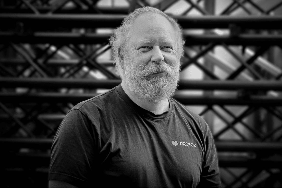 A bearded man with light hair, wearing a dark PROFOX t-shirt, stands in front of a background of metal scaffolding or beams, looking at the camera and smiling slightly. The image is in black and white.