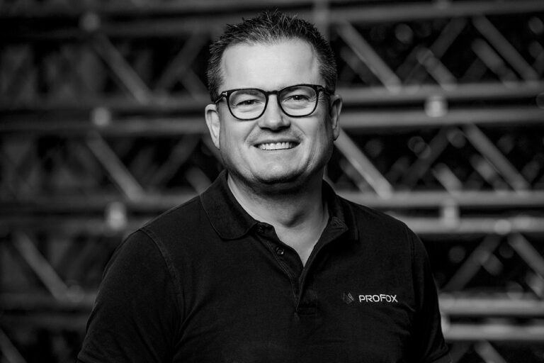 Black and white photo of a smiling man with glasses wearing a collared shirt with PROFIX logo, standing in front of a blurred background with metal shelving or structures.
