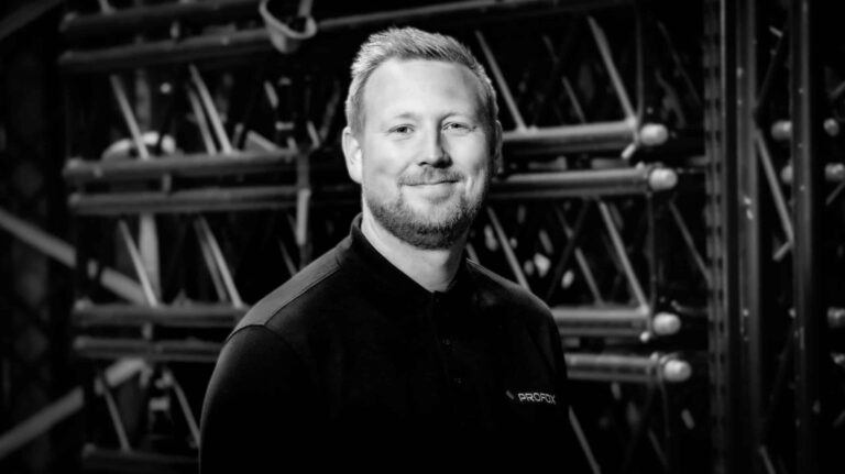 A man with short hair and a beard, wearing a dark polo shirt, smiles at the camera while standing in front of metal scaffolding. The photo is black and white.