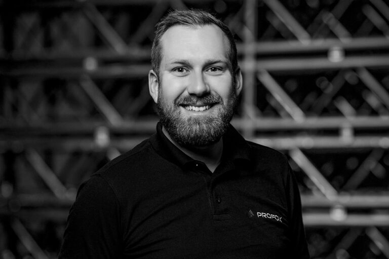Black and white photo of a man with a beard, smiling, wearing a dark collared shirt with a PROGROC logo, standing in front of a blurred industrial-style background.