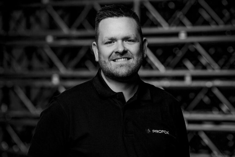 Black and white photo of a smiling man with short hair and a beard, wearing a collared shirt with a PROFOX logo, standing in front of an industrial metal background.