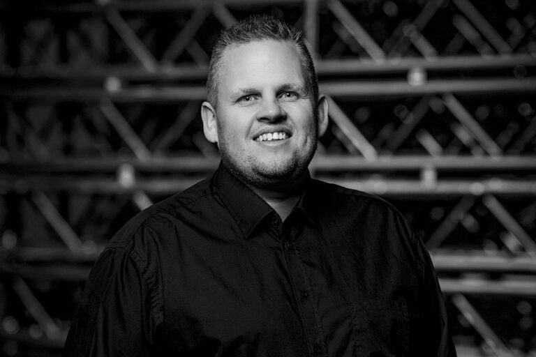 Black and white portrait of a smiling man with short hair, wearing a dark button-up shirt, standing in front of a background with diagonal metal bars.