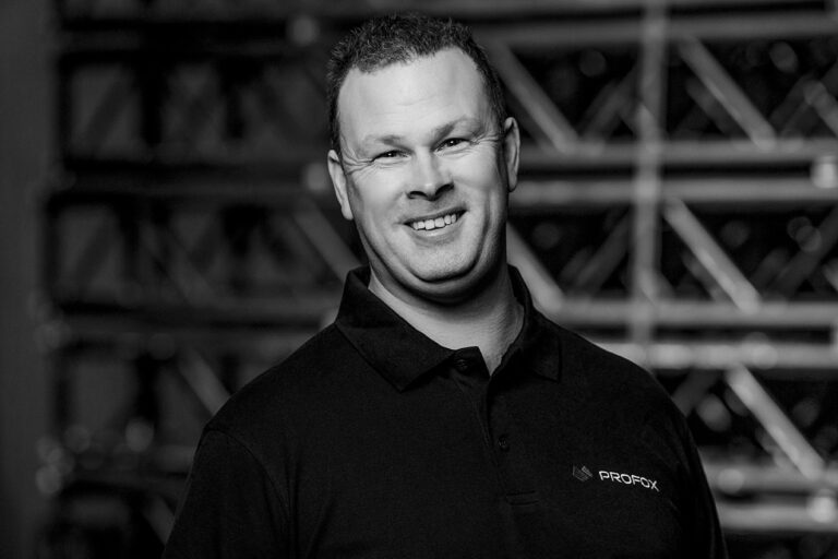 A man with short hair smiles at the camera, wearing a dark polo shirt with a PROFOX logo. The background is blurred with metal structures, and the image is in black and white.