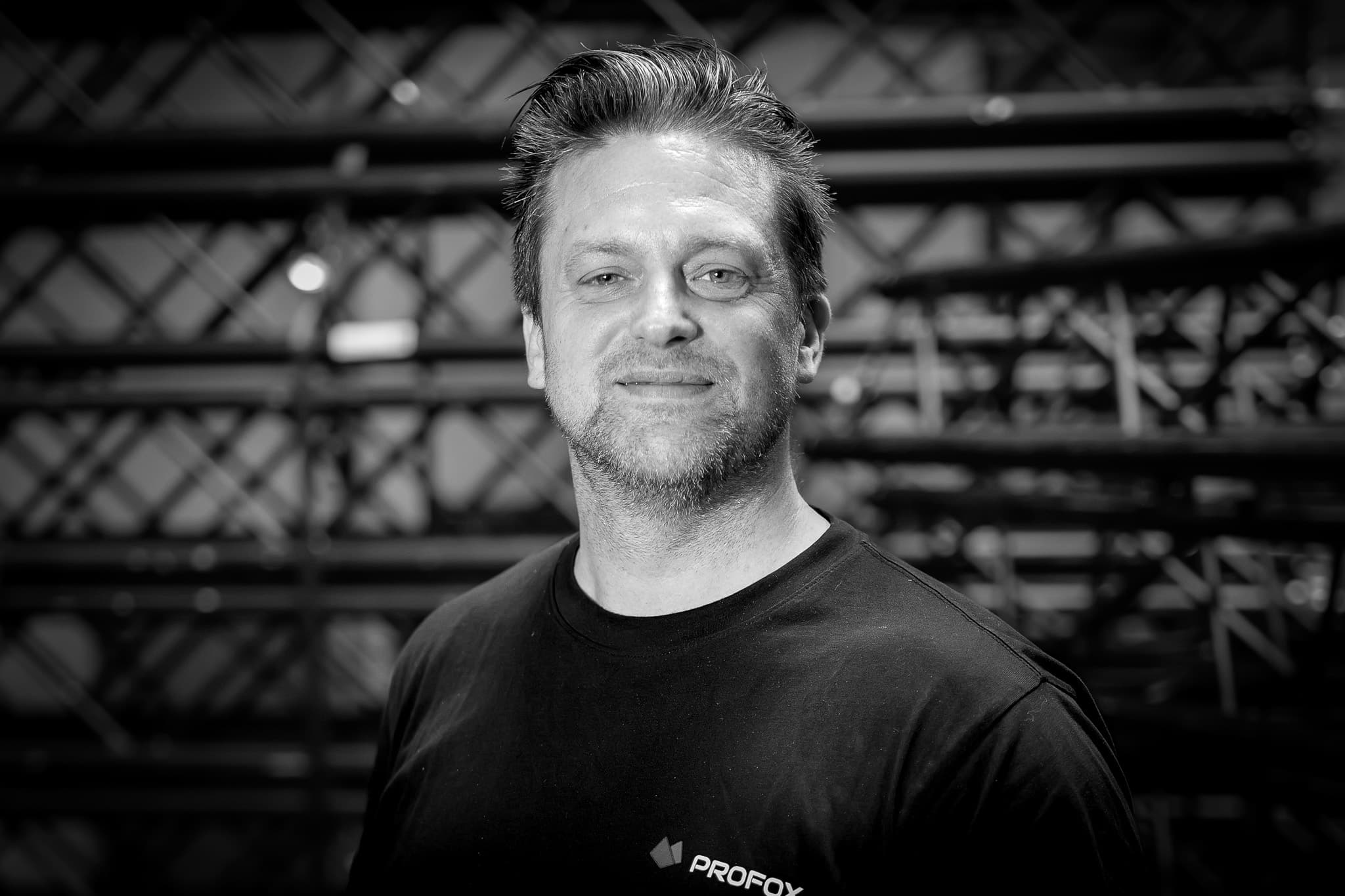 A man with short hair and stubble, wearing a black Profox shirt, stands in front of a blurred background with metal bars, looking at the camera and smiling slightly. The image is in black and white.