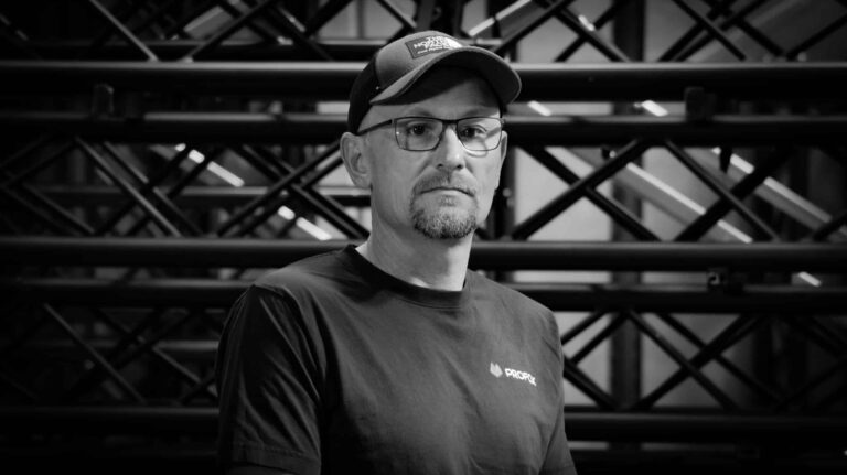 A man wearing glasses, a baseball cap, and a t-shirt stands in front of a background with metal beams and pipes. The image is in black and white.