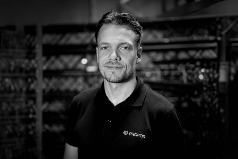 A man with short hair wearing a black PROFOX polo shirt stands in front of industrial shelving. The image is black and white.